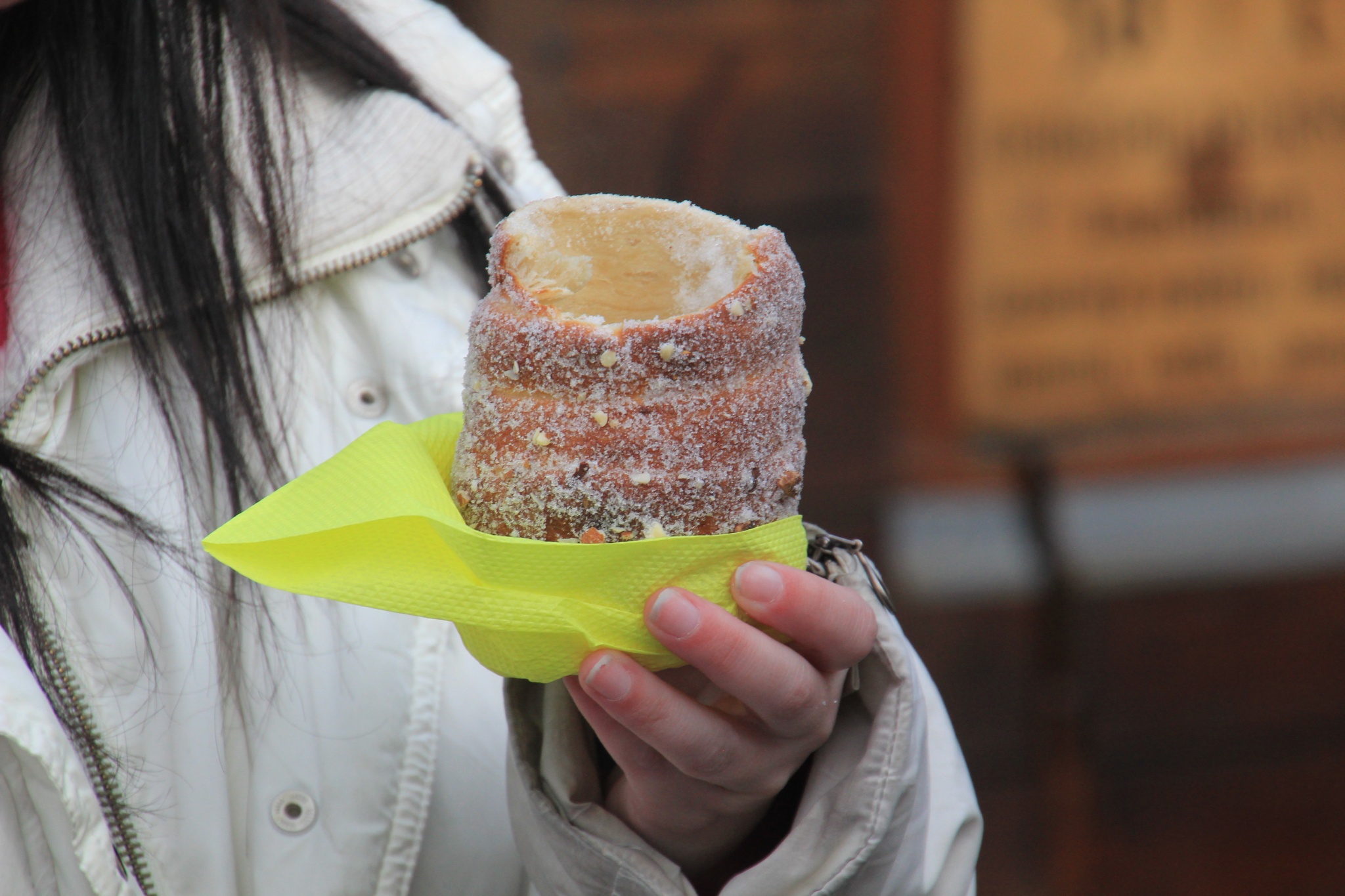 A woman in a white jacket eats a trdelnik in Prague's Old Town Square.