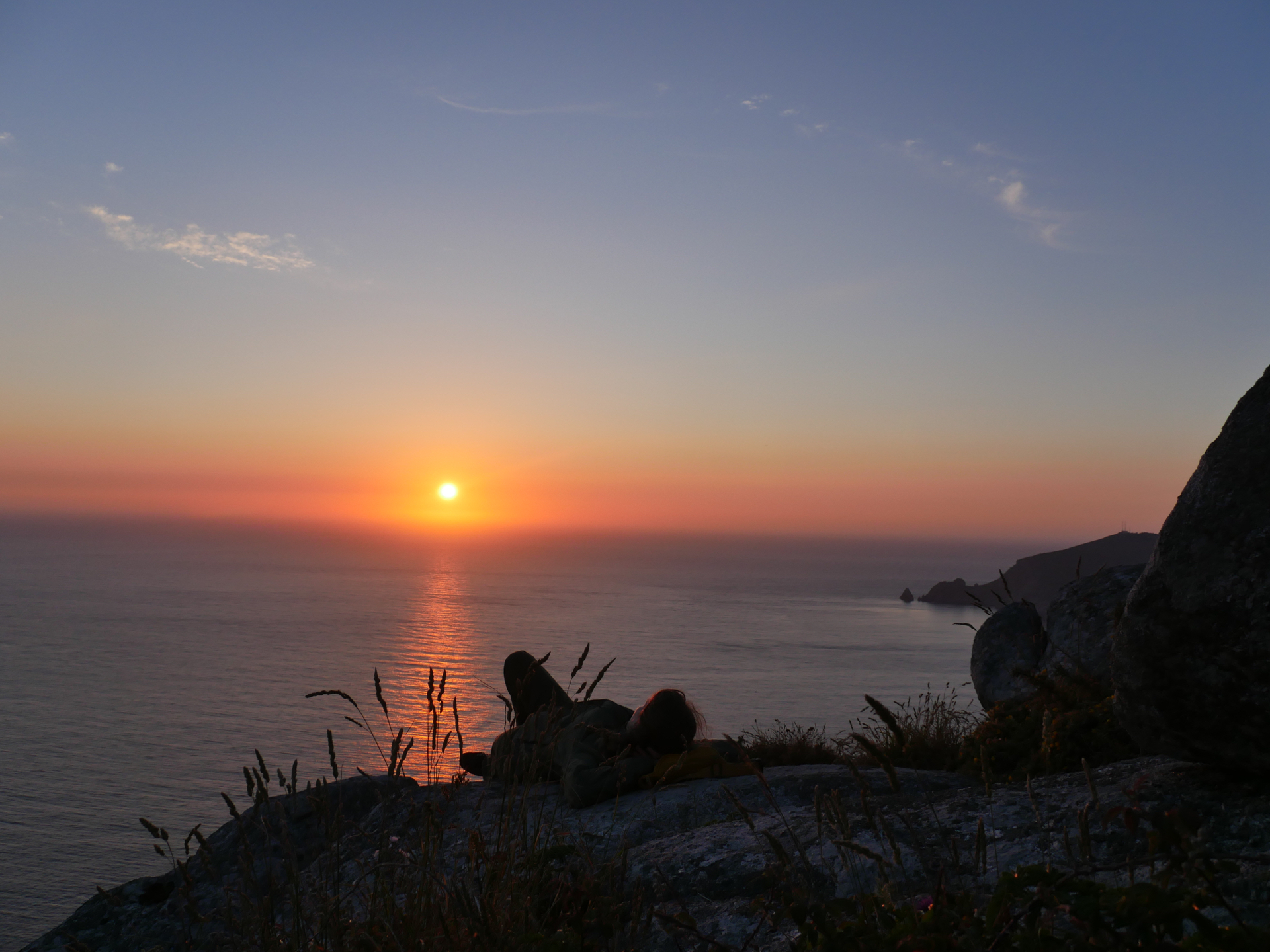 A Camino pilgrim lays on a rock looking at the sunset over the Finisterre peninsula.